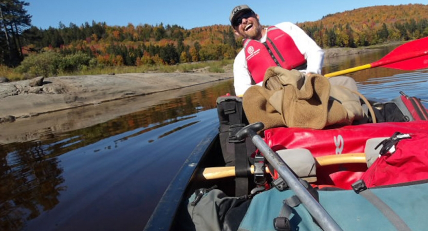 A person sitting in the back of a canoe smiles at the camera. There is fall foliage on the shore behind them.